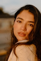 A young woman with long, dark hair gazes softly at the camera. She has a serene expression, with warm light illuminating her features, highlighting her eyes and lips. The background is blurred, with soft, muted colors that suggest an outdoor setting during golden hour.