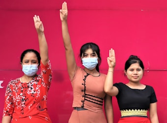 Three women standing against a bright pink background are raising three-finger salutes. The women are wearing masks and various colored dresses, and their expressions appear solemn.