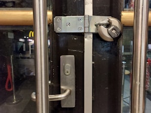 A door with a sturdy silver handle is secured with a large padlock and hasp. The setting appears to be an interior scene of a building, with glass panels on the door, revealing a glimpse of the street outside, featuring visible signage including a McDonald's. The atmosphere is dimly lit, creating a contrast between the interior and external lighting.