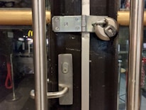 A door with a sturdy silver handle is secured with a large padlock and hasp. The setting appears to be an interior scene of a building, with glass panels on the door, revealing a glimpse of the street outside, featuring visible signage including a McDonald's. The atmosphere is dimly lit, creating a contrast between the interior and external lighting.