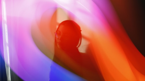 A silhouette of a person, possibly a woman, wearing glasses is seen against a vibrant abstract background of flowing colors including purple, pink, and orange. The scene has an ethereal and dreamy quality.