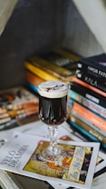 A crystal glass filled with a dark beverage topped with cream sits on a stack of art magazines and books. The glass's ornate design adds elegance, and the magazines include eye-catching designs with artistic illustrations. Books in the background contribute to a cozy, intellectual setting.
