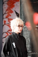 A person with light blonde hair and glasses is wearing a black jacket with a logo featuring a footprint and the letters 'ics'. They are standing in front of an artistic background featuring abstract red branches or coral against a muted backdrop, with a metal or industrial object partially visible on one side.