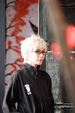A person with light blonde hair and glasses is wearing a black jacket with a logo featuring a footprint and the letters 'ics'. They are standing in front of an artistic background featuring abstract red branches or coral against a muted backdrop, with a metal or industrial object partially visible on one side.