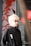 A person with light blonde hair and glasses is wearing a black jacket with a logo featuring a footprint and the letters 'ics'. They are standing in front of an artistic background featuring abstract red branches or coral against a muted backdrop, with a metal or industrial object partially visible on one side.