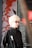 A person with light blonde hair and glasses is wearing a black jacket with a logo featuring a footprint and the letters 'ics'. They are standing in front of an artistic background featuring abstract red branches or coral against a muted backdrop, with a metal or industrial object partially visible on one side.