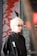 A person with light blonde hair and glasses is wearing a black jacket with a logo featuring a footprint and the letters 'ics'. They are standing in front of an artistic background featuring abstract red branches or coral against a muted backdrop, with a metal or industrial object partially visible on one side.