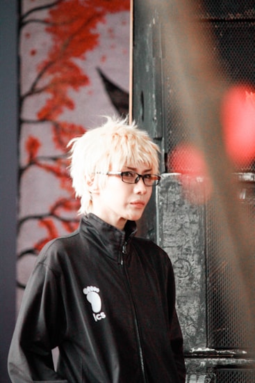 A person with light blonde hair and glasses is wearing a black jacket with a logo featuring a footprint and the letters 'ics'. They are standing in front of an artistic background featuring abstract red branches or coral against a muted backdrop, with a metal or industrial object partially visible on one side.