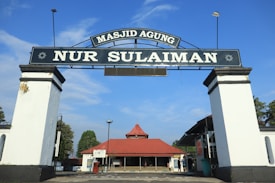 A large archway with the text 'Masjid Agung Nur Sulaiman' stands prominently, leading to a building with a red roof. The sky is blue with few clouds, and there are some trees visible in the background.