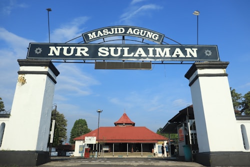 A large archway with the text 'Masjid Agung Nur Sulaiman' stands prominently, leading to a building with a red roof. The sky is blue with few clouds, and there are some trees visible in the background.