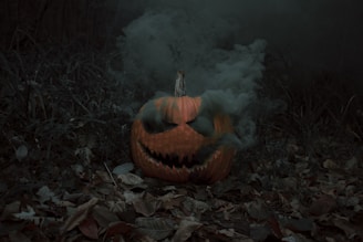 A carved pumpkin with a menacing face emits smoke in a dark, wooded area. Dry autumn leaves and scattered branches cover the ground, enhancing the eerie atmosphere.