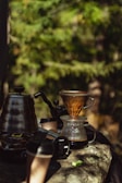 Coffee-making equipment placed on a rock surface in a natural setting. A pour-over coffee setup includes a glass carafe with a coffee filter cone and freshly brewed coffee. Several stainless steel kettles and travel mugs are nearby. The background features blurred green foliage, suggesting an outdoor environment.