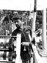 A man wearing a shirt with a logo is working with industrial machinery outdoors. The machinery includes large gears and pipes connected with bolts. The background is a blurred natural setting.