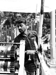 A man wearing a shirt with a logo is working with industrial machinery outdoors. The machinery includes large gears and pipes connected with bolts. The background is a blurred natural setting.