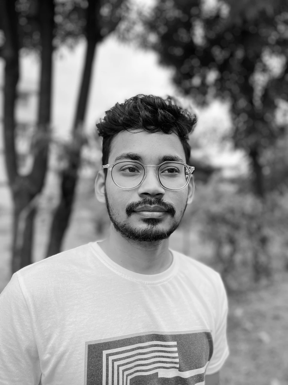 The image shows a man with glasses and a white t-shirt, standing against a backdrop of trees. The man is looking directly at the camera, giving off an air of confidence and self-assurance. His facial hair adds to his distinctive appearance.