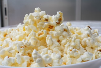 A heap of freshly popped popcorn fills a white bowl, with kernels displaying a light, fluffy texture. The background is softly blurred, emphasizing the popcorn's golden and white hues.