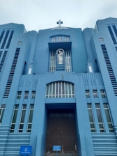 A towering blue building features a prominent cross at the top and a statue of a religious figure in a niche above the entrance. The structure exhibits vertical lines and symmetrical architectural elements, creating a sense of grandeur.