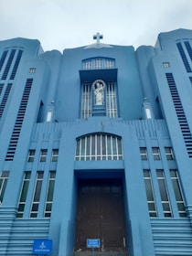 A towering blue building features a prominent cross at the top and a statue of a religious figure in a niche above the entrance. The structure exhibits vertical lines and symmetrical architectural elements, creating a sense of grandeur.