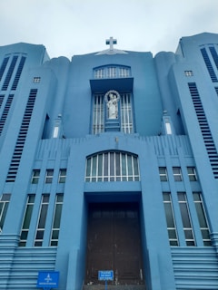 A towering blue building features a prominent cross at the top and a statue of a religious figure in a niche above the entrance. The structure exhibits vertical lines and symmetrical architectural elements, creating a sense of grandeur.