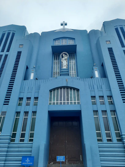 A towering blue building features a prominent cross at the top and a statue of a religious figure in a niche above the entrance. The structure exhibits vertical lines and symmetrical architectural elements, creating a sense of grandeur.