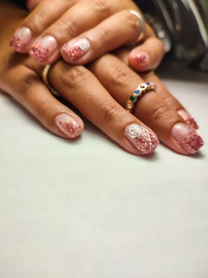 A close-up of hands with a stylish manicure featuring glittery pink nail polish and white rose designs. One hand wears a ring with colorful gemstones, creating an elegant and artistic look.