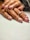A close-up of hands with a stylish manicure featuring glittery pink nail polish and white rose designs. One hand wears a ring with colorful gemstones, creating an elegant and artistic look.