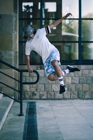 A person wearing a white shirt, blue camouflage shorts, and a black bucket hat is performing a parkour move on a metal railing. The background consists of a glass building and stone walls.