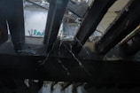 The image features an abandoned or damaged ceiling with wooden beams partially broken and debris visible. Spider webs hang between the beams, and some graffiti can be seen faintly on a distant wall.