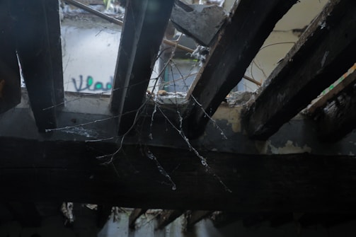 The image features an abandoned or damaged ceiling with wooden beams partially broken and debris visible. Spider webs hang between the beams, and some graffiti can be seen faintly on a distant wall.