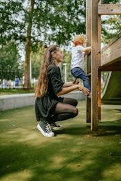 A woman is kneeling on a green playground surface, assisting a young child who is climbing a wooden play structure. The woman is wearing a black dress and sneakers, while the child is in a white shirt, blue pants, and a red cap. Trees and a fenced area are visible in the background.