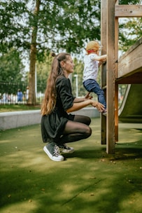 A woman is kneeling on a green playground surface, assisting a young child who is climbing a wooden play structure. The woman is wearing a black dress and sneakers, while the child is in a white shirt, blue pants, and a red cap. Trees and a fenced area are visible in the background.