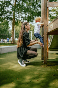 A woman is kneeling on a green playground surface, assisting a young child who is climbing a wooden play structure. The woman is wearing a black dress and sneakers, while the child is in a white shirt, blue pants, and a red cap. Trees and a fenced area are visible in the background.
