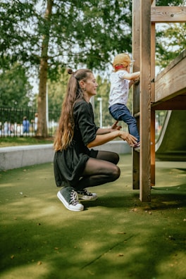 A woman is kneeling on a green playground surface, assisting a young child who is climbing a wooden play structure. The woman is wearing a black dress and sneakers, while the child is in a white shirt, blue pants, and a red cap. Trees and a fenced area are visible in the background.