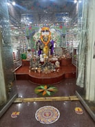 A richly decorated room features a multi-faced deity statue adorned with a vibrant marigold garland. The background is intricately designed with mosaic tiles in bright colors, and various offerings such as fruits and incense are placed at the statue’s base. The floor includes colorful rangoli designs, adding to the festive atmosphere.