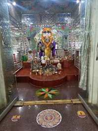 A richly decorated room features a multi-faced deity statue adorned with a vibrant marigold garland. The background is intricately designed with mosaic tiles in bright colors, and various offerings such as fruits and incense are placed at the statue’s base. The floor includes colorful rangoli designs, adding to the festive atmosphere.