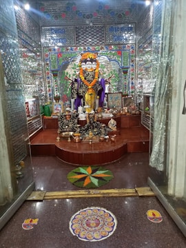 A richly decorated room features a multi-faced deity statue adorned with a vibrant marigold garland. The background is intricately designed with mosaic tiles in bright colors, and various offerings such as fruits and incense are placed at the statue’s base. The floor includes colorful rangoli designs, adding to the festive atmosphere.