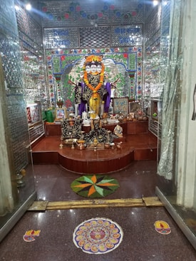 A richly decorated room features a multi-faced deity statue adorned with a vibrant marigold garland. The background is intricately designed with mosaic tiles in bright colors, and various offerings such as fruits and incense are placed at the statue’s base. The floor includes colorful rangoli designs, adding to the festive atmosphere.