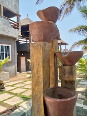 A set of tiered, rustic stone or clay water features, positioned in an outdoor setting near a concrete building with a thatched roof. Palm trees are visible, casting shadows and adding a tropical vibe. The ground is landscaped with stone tiles and grass.