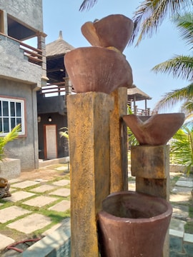 A set of tiered, rustic stone or clay water features, positioned in an outdoor setting near a concrete building with a thatched roof. Palm trees are visible, casting shadows and adding a tropical vibe. The ground is landscaped with stone tiles and grass.