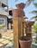 A set of tiered, rustic stone or clay water features, positioned in an outdoor setting near a concrete building with a thatched roof. Palm trees are visible, casting shadows and adding a tropical vibe. The ground is landscaped with stone tiles and grass.
