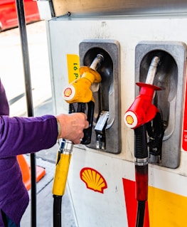 A person is refueling at a gas pump station. Two fuel nozzles are visible, one red and one yellow, bearing the Shell logo. The individual is wearing a purple sleeve and holding the yellow nozzle. A traffic cone and part of a red vehicle are visible in the background.
