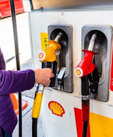 A person is refueling at a gas pump station. Two fuel nozzles are visible, one red and one yellow, bearing the Shell logo. The individual is wearing a purple sleeve and holding the yellow nozzle. A traffic cone and part of a red vehicle are visible in the background.