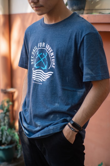 A person wearing a blue t-shirt with the text 'Set Sail for Adventure' and a graphic of two crossed anchors. The person has one hand in their pocket and is wearing a black wristband or watch. The background features warm tones with some plants.