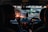 The interior of a car with two people seated in the front, likely the driver and a passenger. The dashboard is illuminated, and the outside view through the windshield shows city lights and blurred traffic. The ambient lighting suggests it is evening or nighttime.