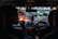 The interior of a car with two people seated in the front, likely the driver and a passenger. The dashboard is illuminated, and the outside view through the windshield shows city lights and blurred traffic. The ambient lighting suggests it is evening or nighttime.