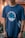 A person is wearing a navy blue t-shirt with a nautical theme design. The shirt features crossed anchors and waves, alongside the text 'Set Sail for Adventure'. The background includes blurred greenery and a warm-toned wall.