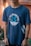 A person is wearing a navy blue t-shirt with a nautical theme design. The shirt features crossed anchors and waves, alongside the text 'Set Sail for Adventure'. The background includes blurred greenery and a warm-toned wall.