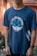 A person is wearing a navy blue t-shirt with a nautical theme design. The shirt features crossed anchors and waves, alongside the text 'Set Sail for Adventure'. The background includes blurred greenery and a warm-toned wall.