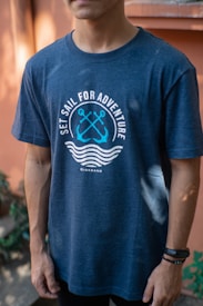 A person is wearing a navy blue t-shirt with a nautical theme design. The shirt features crossed anchors and waves, alongside the text 'Set Sail for Adventure'. The background includes blurred greenery and a warm-toned wall.