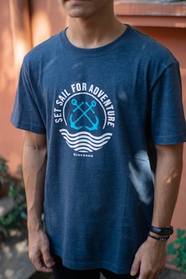 A person is wearing a navy blue t-shirt with a nautical theme design. The shirt features crossed anchors and waves, alongside the text 'Set Sail for Adventure'. The background includes blurred greenery and a warm-toned wall.